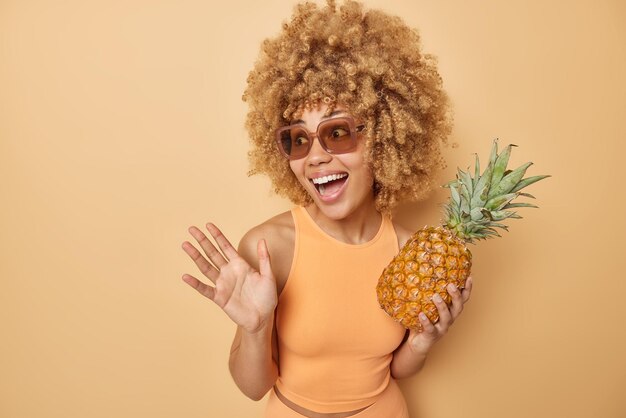 Una foto horizontal de una mujer alegre y despreocupada con el pelo rubio rizado sostiene una piña fresca que mantiene la palma levantada se ríe alegremente usa gafas de sol de moda, una camiseta informal aislada sobre un fondo beige