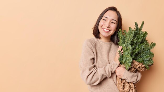 La foto horizontal de una mujer asiática feliz con una sonrisa dentuda abraza un ramo de ramas de abeto verde sueña con el próximo Año Nuevo y Navidad aislado sobre una pared beige con espacio en blanco para la promoción