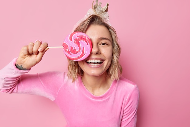 La foto horizontal de una mujer bonita joven y positiva con el peinado cubre los ojos con un caramelo redondo lleva corona y ropa festiva sonríe ampliamente muestra dientes blancos aislados sobre un fondo rosa