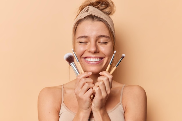 La foto horizontal de una mujer complacida cierra los ojos, sonríe ampliamente, sostiene cepillos cosméticos para aplicar maquillaje, usa una camiseta informal con banda para la cabeza aislada sobre un fondo beige. Concepto de cuidado de la piel facial.