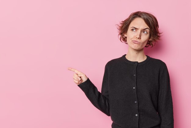 La foto horizontal de una mujer disgustada con el pelo corto levanta las cejas, se enfurruña, señala con el dedo índice en el espacio en blanco y anuncia algo aislado sobre un fondo rosa. Mira esta promoción.