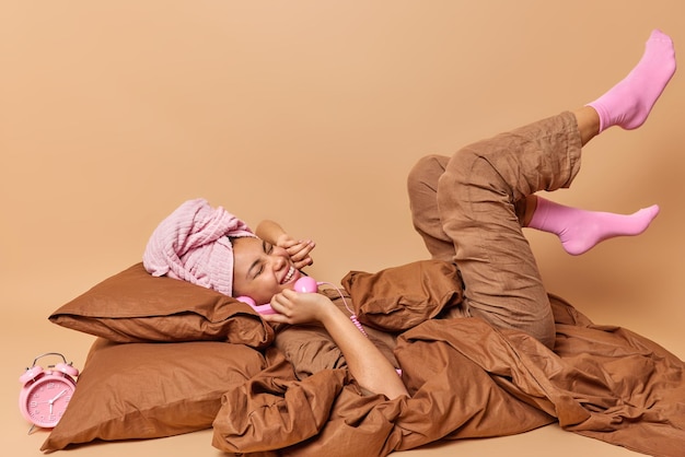 Una foto horizontal de una mujer feliz y perezosa se queda en la cama durante mucho tiempo y tiene una conversación telefónica que mantiene el auricular retro cerca de la oreja vestido con cómodas poses de pijama en el dormitorio con fondo marrón