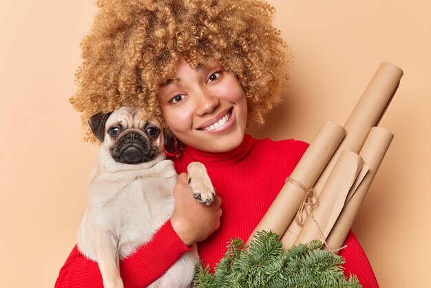 La foto horizontal de una mujer feliz se ve con una expresión amistosa que abraza las sonrisas favoritas de las mascotas sostiene agradablemente papel decorativo y ramas de abeto verde viste poloneck rojo aislado sobre una pared beige