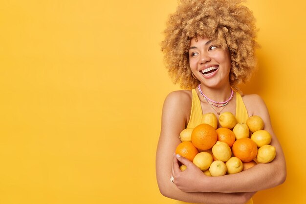 La foto horizontal de una mujer joven y bonita abraza naranjas y frutas frescas y jugosas tiene una expresión alegre sonrisa con dientes en la cara aislada sobre el espacio de copia de fondo amarillo para su contenido promocional