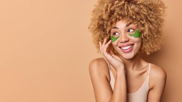 La foto horizontal de una mujer joven y bonita con el pelo rizado y tupido mira hacia otro lado y tiene una expresión facial alegre que se pone parches verdes hidratantes debajo de los ojos para quitar las bolsas y las arrugas y parece sonreír