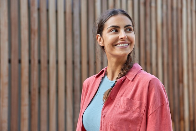 Una foto horizontal de una mujer muy sonriente con el pelo oscuro peinado con coleta vestida con una camisa rosa informal enfocada con expresión alegre pasea al aire libre durante el día contra el fondo de madera