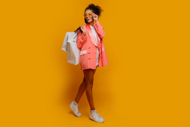 Foto integral de la elegante chica estadounidense con piel oscura en zapatillas blancas de pie con bolsas de compras sobre fondo amarillo.