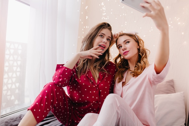 Foto interior de adorable chica rubia con teléfono para selfie con amiga. Entusiasta dama morena en camisón de algodón rojo posando con su hermana.