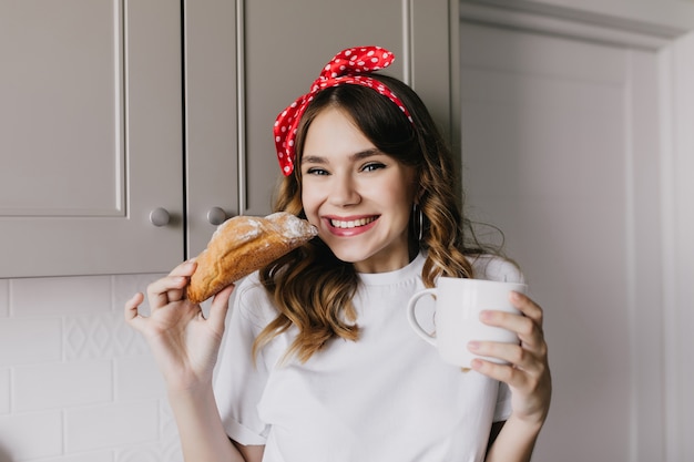 Foto interior de una chica maravillosa comiendo croissant dulce. Retrato de increíble dama europea divirtiéndose durante el almuerzo.