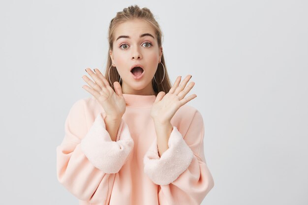 Foto interior de una joven excitada con el pelo rubio con una mirada sorprendida, exclamando, manteniendo la boca abierta y las manos sorprendidas con su inesperada victoria o presente.