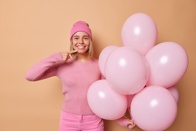 La foto interior de una joven feliz pasa tiempo libre en los puntos de la despedida de soltera en su sonrisa con dientes sostiene un gran montón de globos de helio, usa un jersey de sombrero y pantalones aislados sobre un fondo marrón