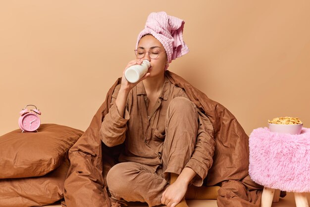 La foto interior de una joven vestida con una cómoda toalla de pijama envuelta en la cabeza bebe leche fresca posada bajo una manta en la cama cerca de almohadas tiene un desayuno rápido aislado sobre un fondo beige.