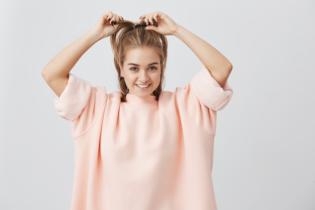 Foto interior de una mujer hermosa, encantadora y joven que se arregla el cabello rubio y liso, sonriendo ampliamente, demostrando sus dientes blancos perfectos. Encantadora niña tierna vestida de rosa.