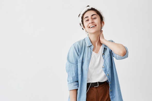 La foto de interior de una mujer joven con cabello oscuro y ondulado en moño, usa camisa vaquera y pantalones marrones, con la mano en el cuello, usa un teléfono móvil moderno para entretenerse, escucha su música favorita con auriculares