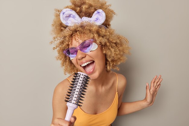 Foto gratuita la foto interior de una mujer rubia alegre sostiene un peine cerca de la boca como si el micrófono se sintiera muy feliz aplica parches de belleza debajo de los ojos usa una diadema de gafas de sol moradas y una camiseta aislada en una pared gris