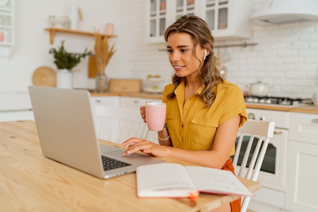 Foto interior de mujer rubia sonriente usando laptop durante el desayuno en su moderna cocina ligera.