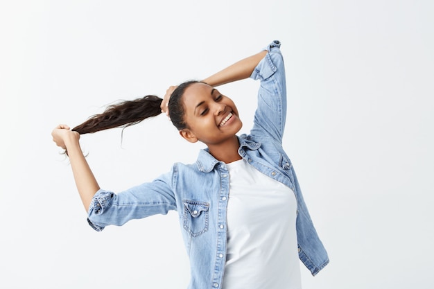 Foto interior de una niña afroamericana con el pelo largo y ondulado recogido en una cola de caballo y piel oscura vestida con una camisa de mezclilla posando en el interior, jugando con su cabello.