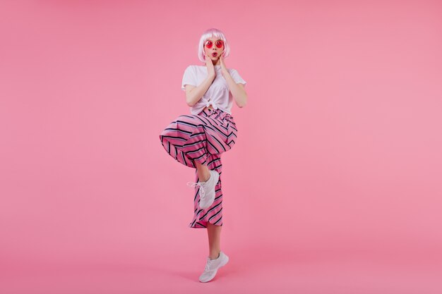 Foto interior de niña bonita sorprendida en rosa periwig divertida posando en la pared rosa. refinada dama caucásica en traje de moda bailando y expresando emociones de sorpresa