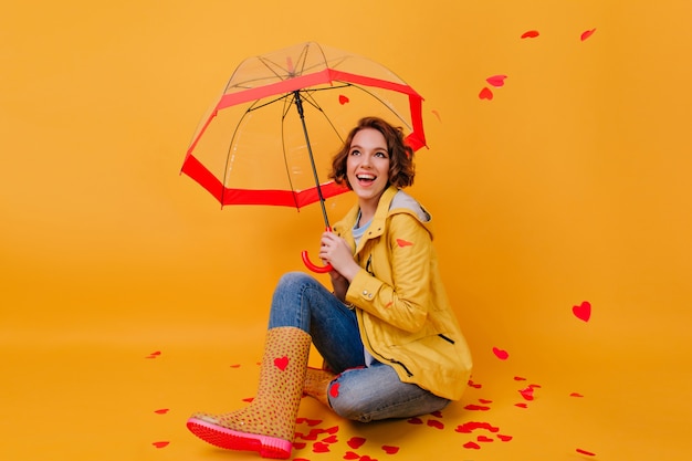 Foto interior de niña en zapatos de goma de moda riendo bajo el paraguas. Foto de estudio de dama extasiada jugando en el día de San Valentín.