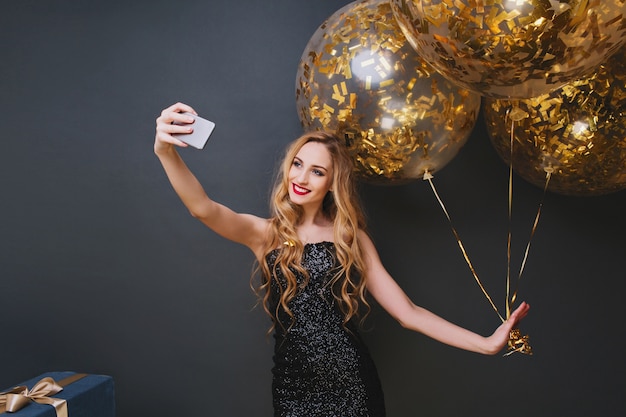 Foto interior de una romántica cumpleañera rubia haciendo selfie. Sonriente mujer caucásica rizada escalofriante en la fiesta con globos y regalos.