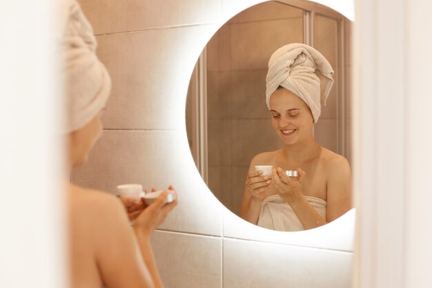 Foto en interiores de una mujer sonriente que hidrata la piel después de la ducha, sostiene la crema en las manos, expresa emociones felices, se envuelve en una toalla y realiza procedimientos de spa en casa.