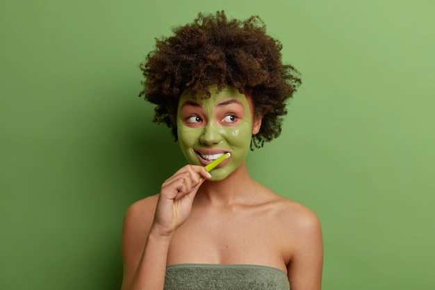 Foto de joven afroamericana aplica mascarilla verde cepillos dientes utiliza cepillo de dientes