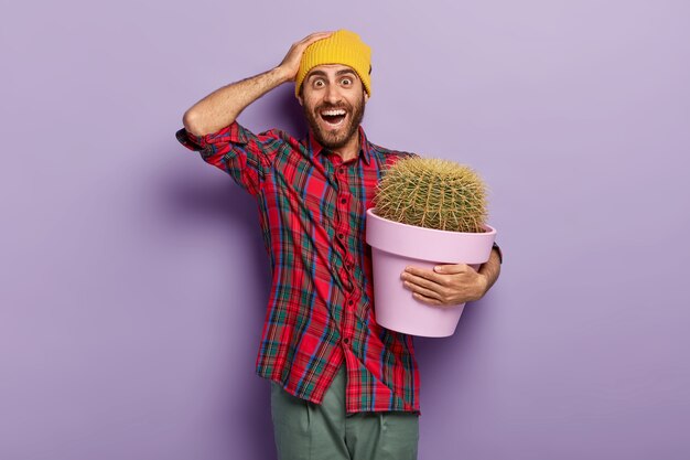 Foto de un joven alegre sin afeitar que mantiene la mano en la cabeza, lleva una maceta de planta verde, recibe cactus con espinas espinosas como presente, usa un sombrero amarillo y una camisa roja trenzada, se preocupa por la planta en maceta