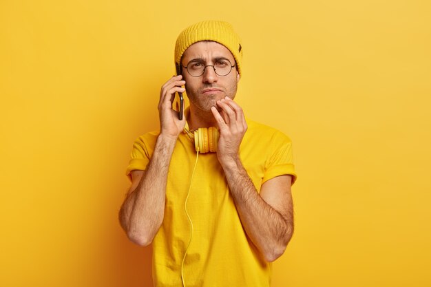 Foto de joven atento serio tiene conversación telefónica, mantiene el teléfono móvil moderno cerca del oído, tiene auriculares