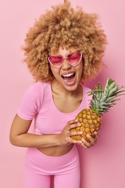 Foto de una joven de cabello rizado muy contenta que exclama de alegría usa gafas de sol de moda vestidas con ropa deportiva sostiene piña fresca llena de vitaminas aisladas sobre fondo rosa Fruta tropical