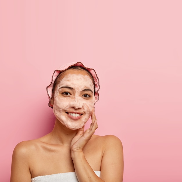 Foto de una joven china que disfruta de la purificación de la piel de la cara, se lava con jabón, toca la mejilla, se ve con una sonrisa, limpia los poros, usa gorro de baño, posa en el interior, copia espacio en la pared rosa