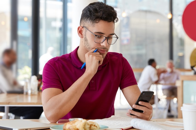 Foto de joven concentrado con peinado moderno, cerda pequeña, lee noticias del mensaje de ingresos, sostiene el teléfono inteligente en una mano y el bolígrafo en la otra, hace registros, se prepara para una reunión de negocios