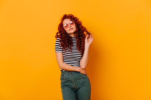 Foto de joven coqueta adolescente en jeans de mamá y camiseta a rayas tocando sus rizos en el espacio naranja.