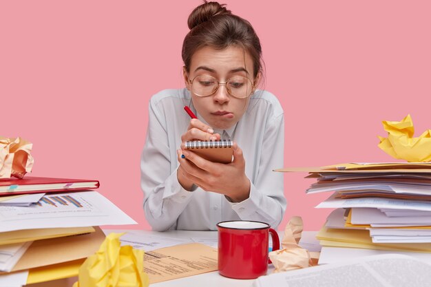 Foto de joven curiosa inteligente escribe en el bloc de notas, sostiene la pluma tiene mirada atenta, usa gafas grandes y camisa blanca, hace registros