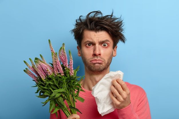 Foto de un joven enfermo infeliz que tiene problemas de salud, se frota la nariz con un pañuelo de papel, sufre de estornudos y ojos llorosos, tiene alergia a la planta de primavera estacional, hipersensibilidad a la floración