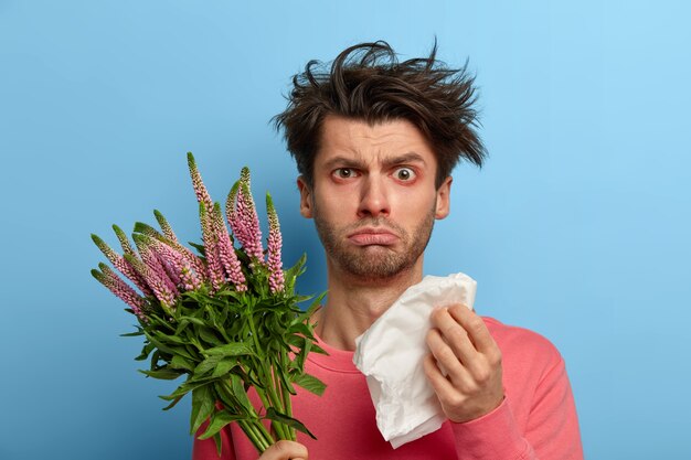 Foto de un joven enfermo infeliz que tiene problemas de salud, se frota la nariz con un pañuelo de papel, sufre de estornudos y ojos llorosos, tiene alergia a la planta de primavera estacional, hipersensibilidad a la floración