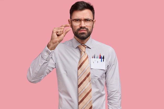 Foto de un joven escupuloso con barba espesa, muestra algo pequeño con la mano, demuestra préstamo pequeño, usa anteojos grandes