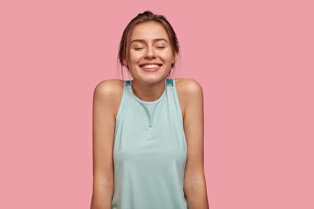 Foto de una joven europea satisfecha con una amplia sonrisa agradable, mantiene los ojos cerrados, sueña con algo agradable, muestra los hombros desnudos, vestida con un chaleco informal, aislada sobre una pared rosa