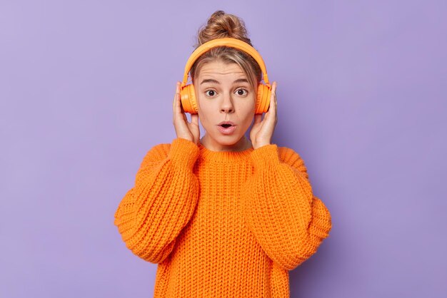 Foto de una joven europea sorprendida que aguanta el aliento, no puede creer que sus propios ojos usen un suéter suelto de punto, escuchen música a través de auriculares inalámbricos, posa en el interior contra el fondo púrpura, el concepto Omg