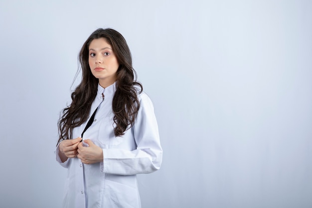 Foto de joven médico en bata blanca de pie sobre una pared blanca.