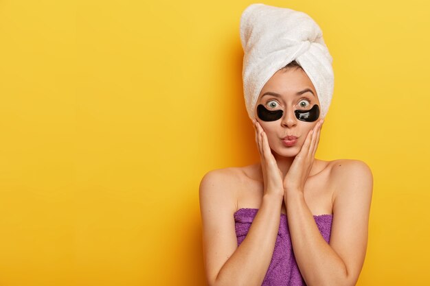 Foto de una joven modelo sorprendida que toca las mejillas, mantiene los labios redondeados, aplica parches negros debajo de los ojos, reduce la superficie de la piel, usa una toalla envuelta