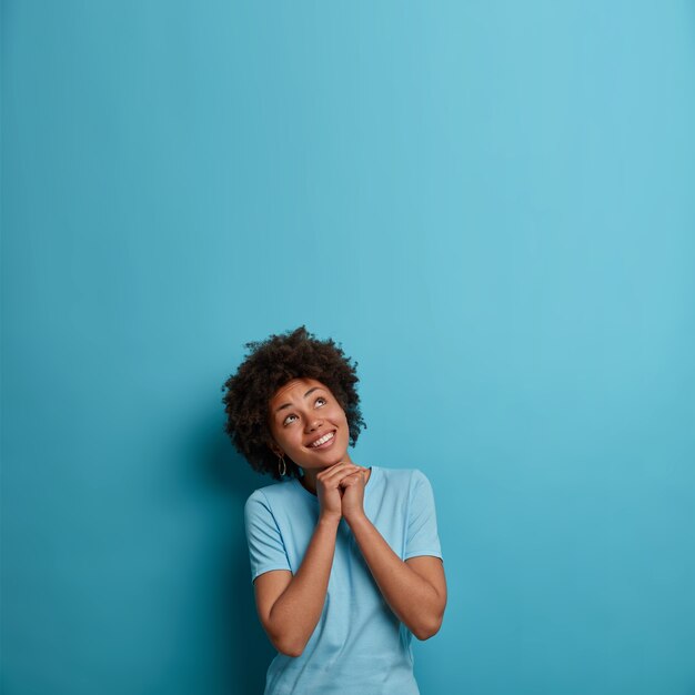 Foto de una joven optimista que mantiene las manos juntas debajo de la barbilla, mira hacia arriba, feliz de ver descuentos agradables, cree y espera algo mejor, usa una camiseta azul informal, espacio vacío hacia arriba