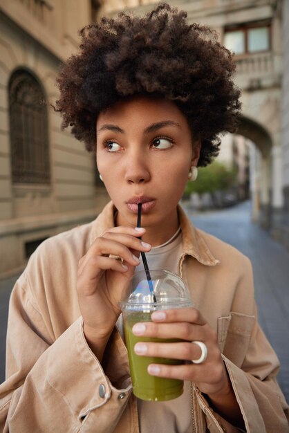 La foto de una joven pensativa bebe jugo vegetariano o batido de vegetales verdes usa paja enfocada en poses en un entorno urbano cuida la salud mantiene la dieta nutrición paseos al aire libre