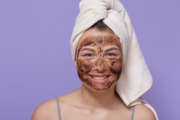 Foto de joven sonriente aplicando máscara cosmética marrón en la cara, limpiando su piel en casa