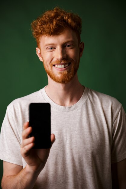 Foto de joven sonriente barbudo pelirrojo con camiseta blanca hace una foto en el teléfono inteligente móvil