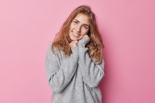 Foto de una joven tierna y de buen aspecto que inclina la cabeza mantiene las manos cerca de la cara sonríe suavemente tiene un aspecto agradable y lleva un puente gris suave aislado sobre un fondo rosa. Lindo modelo femenino positivo