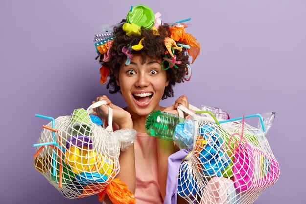 Foto de una joven voluntaria feliz que lleva bolsas con basura, se ve alegre, feliz de hacer cosas buenas por el medio ambiente y el planeta donde vive, aislada sobre una pared púrpura.