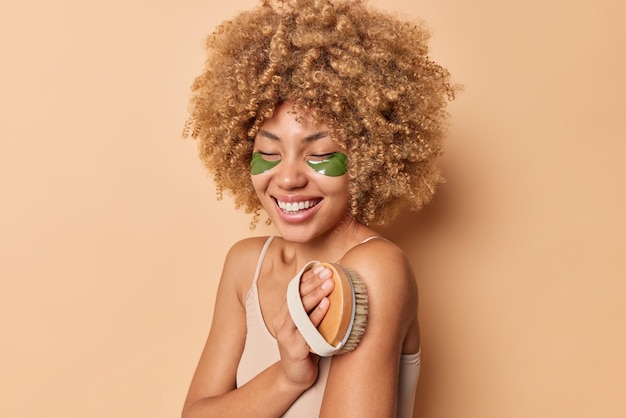 La foto lateral de una mujer joven con el cuerpo de cepillos de pelo rizado y tupido con un cepillo de madera aplica parches de hidrogel verde debajo de los ojos posa contra el fondo marrón del estudio. Concepto de cuidado del cuerpo de anuncio de belleza