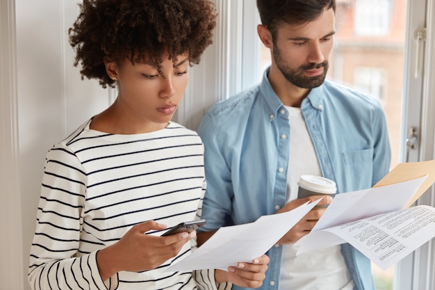 Foto de una lista de verificación de estudios de mujer negra seria, usa calculadora en el teléfono móvil, tipo sin afeitar con cerdas sostiene café, aprende auditoría de trabajo, prepara informe financiero anual, verifica inspección