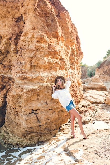 Foto de longitud completa de una chica bonita rubia con el pelo largo posando para la cámara en la playa sobre fondo de roca.