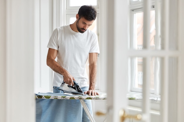 Foto de marido responsable o hombre soltero ocupado con el trabajo de la casa, plancha su camisa por la mañana en la mesa de planchado antes del trabajo